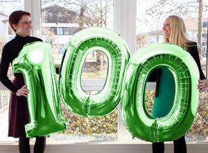 Avison Erfolgsgeschichte: Zwei Mitarbeiterinnen halten die Zahl 100 als grüne Luftballons